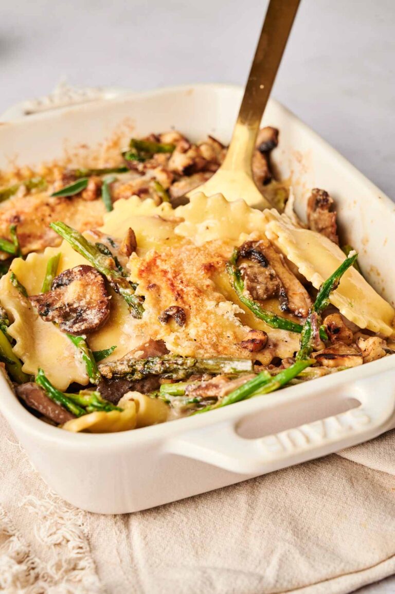 A ravioli casserole with mushrooms, green beans, and a golden cheese crust, all nestled in a rectangular white baking dish with a serving spoon.