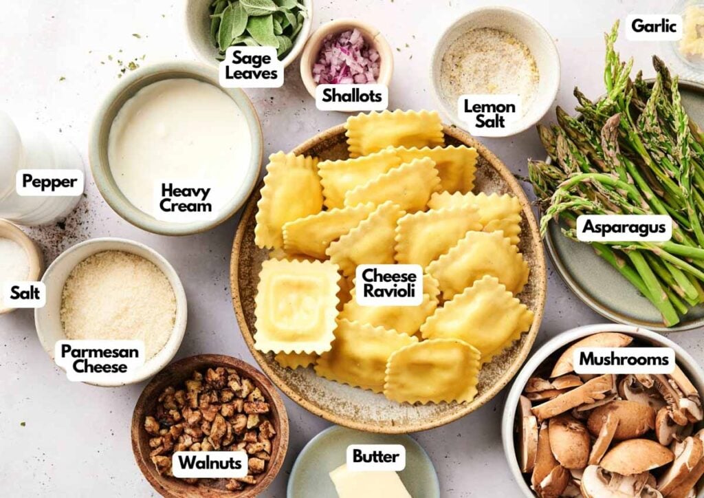 A bowl of cheese ravioli casserole is surrounded by small dishes containing asparagus, mushrooms, walnuts, butter, heavy cream, sage leaves, shallots, garlic, lemon salt, parmesan, salt, and pepper.