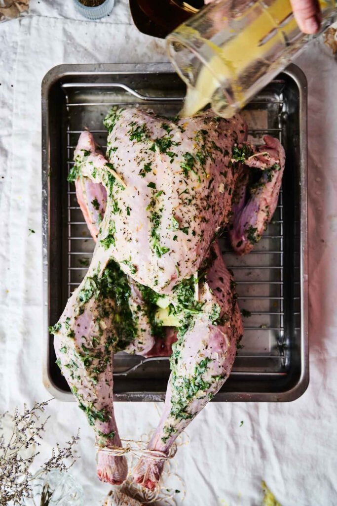 Raw turkey seasoned with herbs on a roasting rack, with a hand pouring liquid marinade over it.