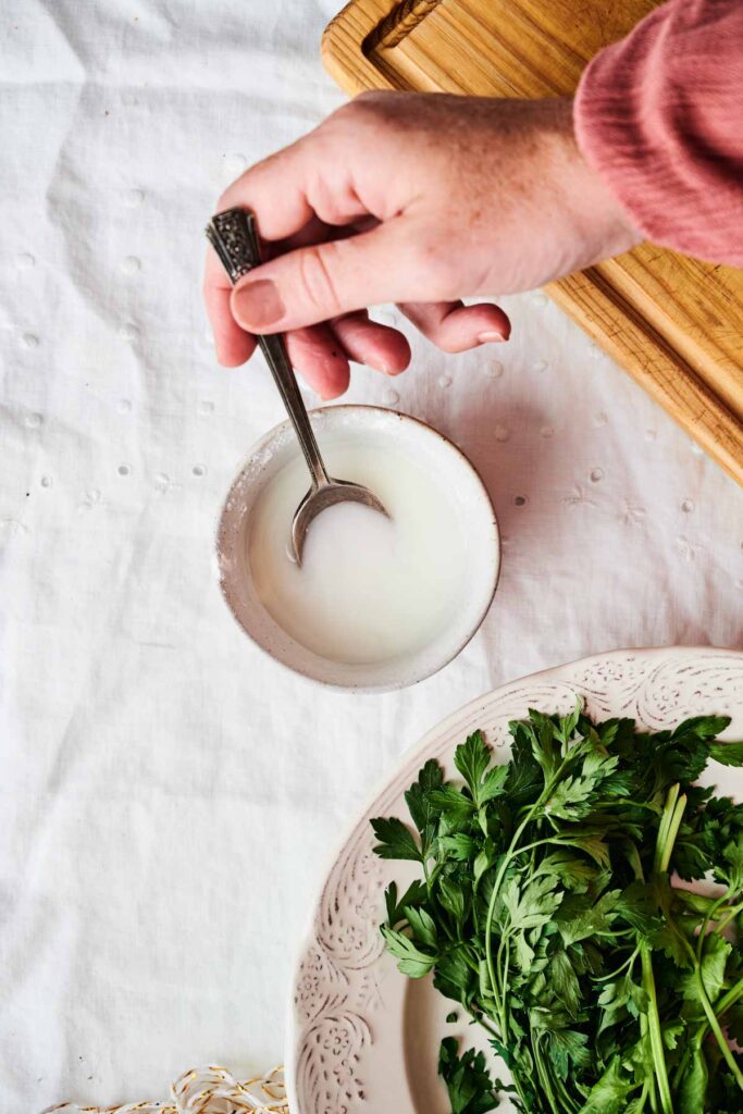 A hand stirring a small bowl of liquid with a spoon, next to a plate of fresh parsley on a white tablecloth.