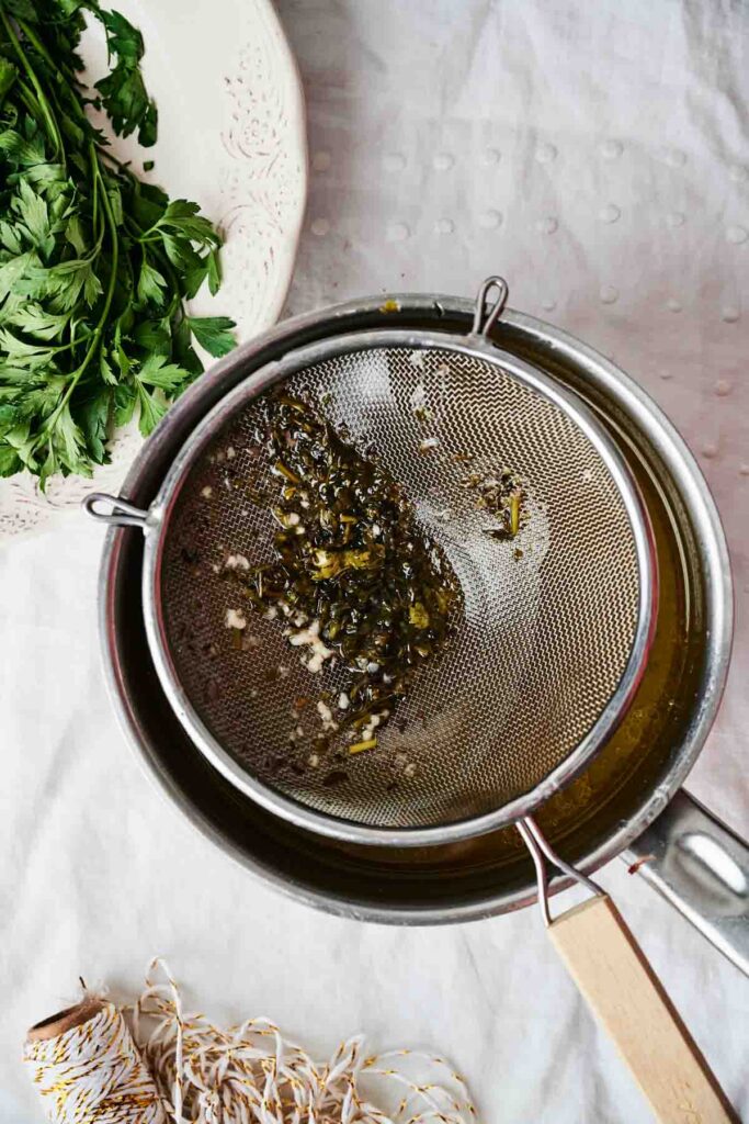Herbs in a mesh strainer over a saucepan, with fresh parsley on a plate and string nearby on a white textured surface.