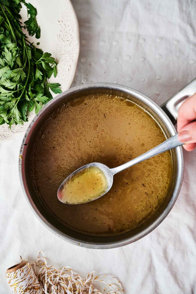 A hand holds a spoon of broth above a pot on a table, with fresh parsley and twine nearby.