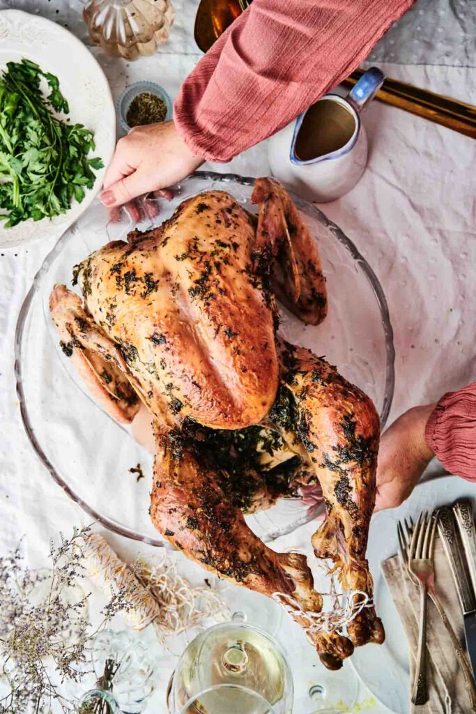 A whole roasted turkey garnished with herbs on a platter, surrounded by greens and a jug, with a person's hands adjusting it on a dining table.