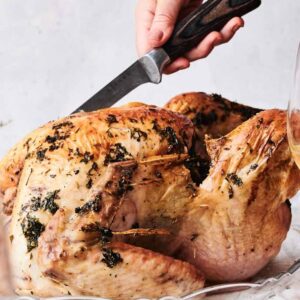 A person carving a roasted turkey on a glass platter, surrounded by wine glasses and a set table with white linens and dried flowers.