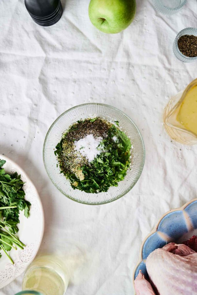 A glass bowl on a table contains chopped herbs and seasoning. Nearby are parsley, an apple, a container of spices, and a dish with raw poultry.