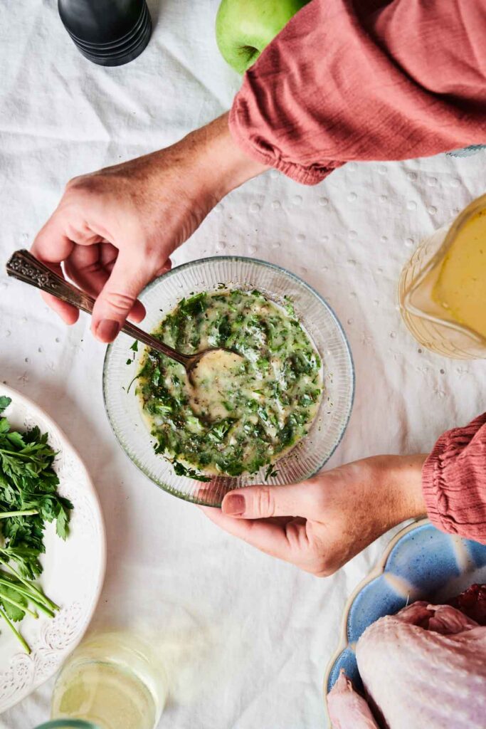 Hands preparing a green herb sauce in a glass bowl, surrounded by raw chicken, parsley, an apple, and a pitcher on a white tablecloth.