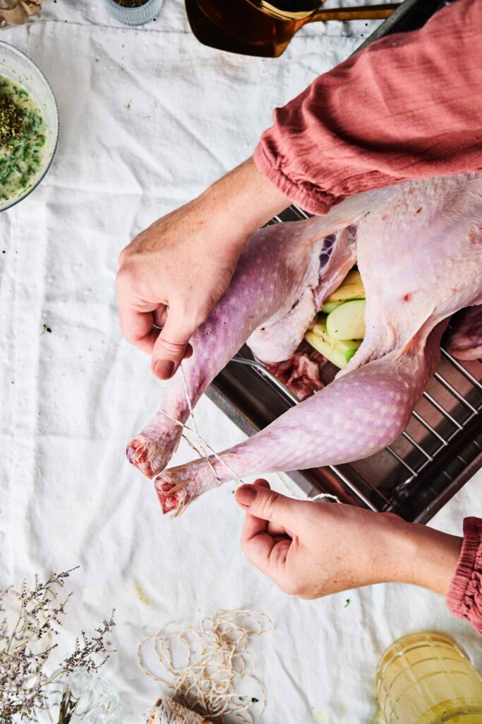 Person trussing a raw turkey with string on a white surface, preparing it for cooking.