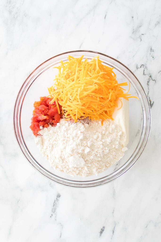 A bowl containing shredded cheese, diced tomatoes, flour, and a white block of cream cheese sits on a marble surface, perfect for whipping up Rotel-infused dishes or savory sausage balls.