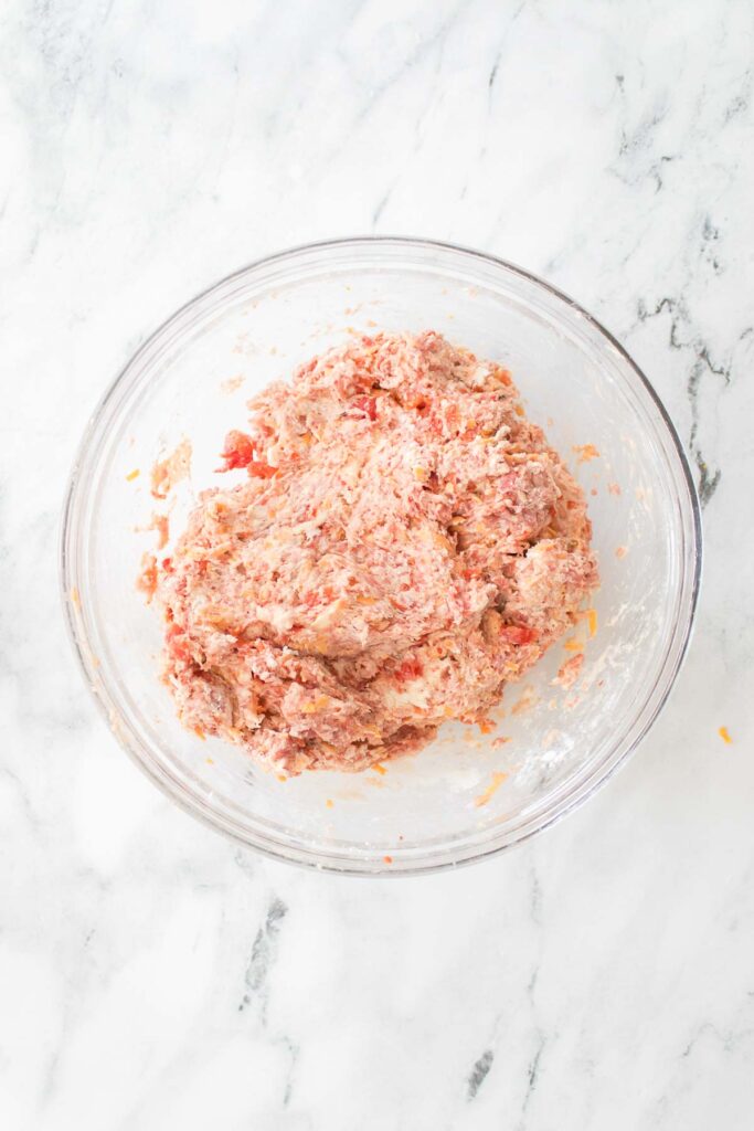 A glass bowl on a marble surface contains ground meat mixed with spices, ready to transform into savory sausage balls.