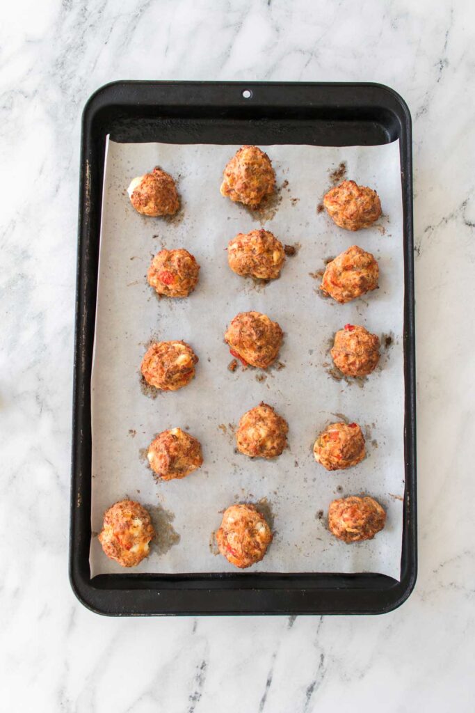 A baking tray with fifteen evenly spaced Rotel cream cheese sausage balls rests on parchment paper, placed on a marble countertop.
