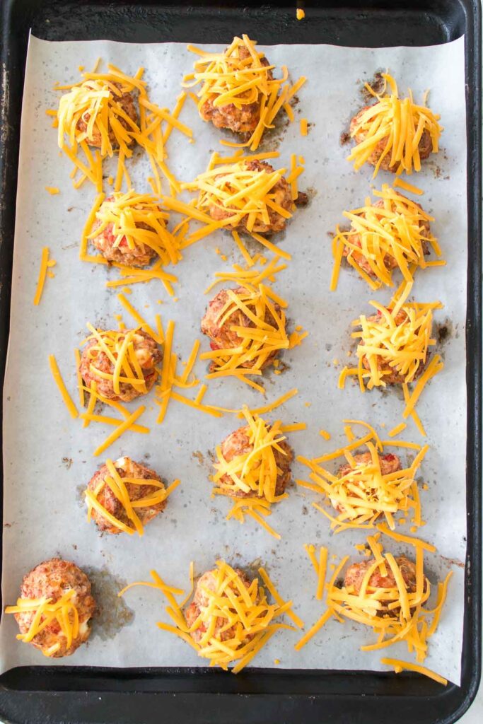Sausage balls made from ground meat are enhanced with Rotel and cream cheese, topped with shredded cheddar, and arranged on a parchment-lined baking tray.