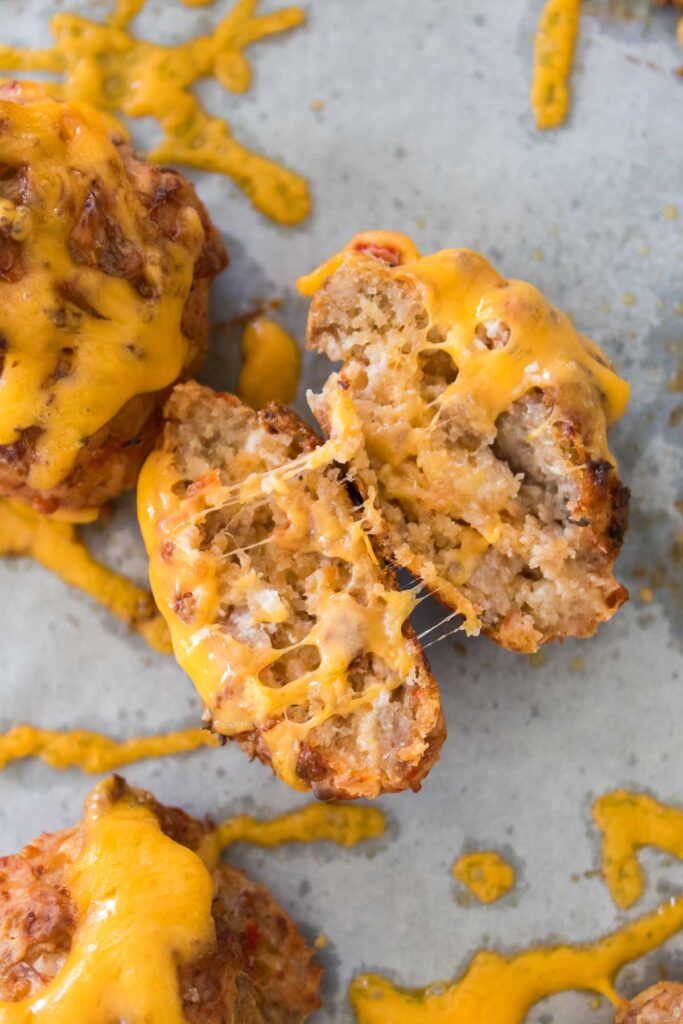Muffin-shaped baked goods with melted cheese, split in half to reveal a crumbly texture, sit on a grey surface with cheese drips. Their savory essence hints at the comforting flavors found in classic Rotel Cream Cheese Sausage Balls.