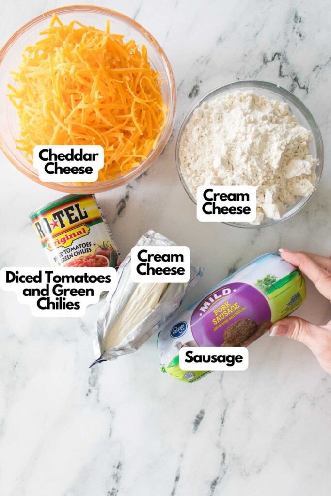 Ingredients for Rotel Cream Cheese Sausage Balls on a marble countertop: a bowl of shredded cheddar cheese, two packages of cream cheese, canned diced tomatoes with green chilies, and a package of savory sausage.