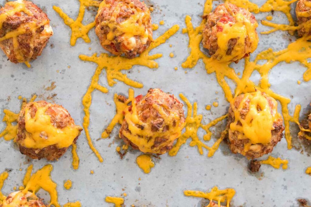 Rotel cream cheese sausage balls with visible cheese drips rest enticingly on a baking sheet, blending classic flavors with a spicy twist.