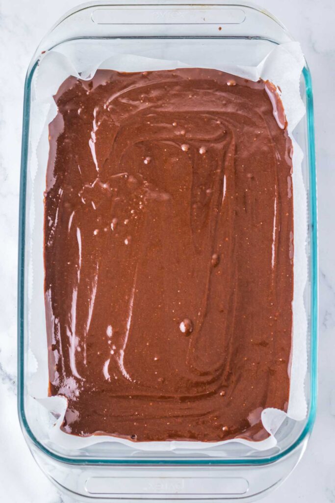 A glass baking dish filled with Pink Snoball Brownie batter sits enticingly on parchment paper, gracefully poised atop a sleek marble surface.