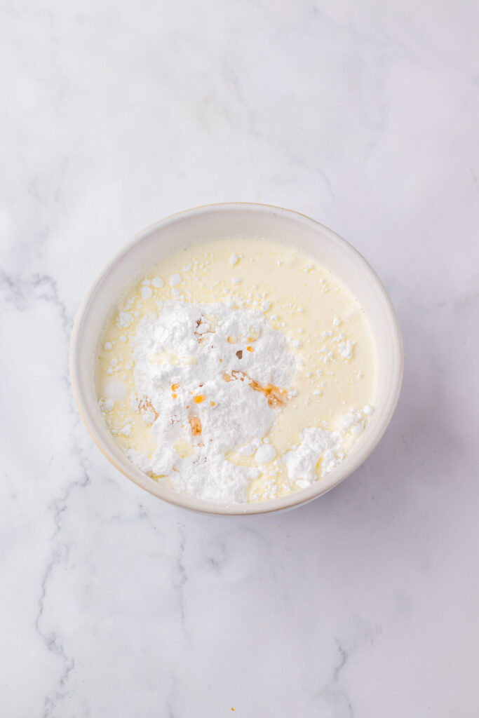 A bowl with cream, powdered sugar, and vanilla extract on a marble surface awaits to create the perfect frosting for a copycat Starbucks Red Velvet Loaf.