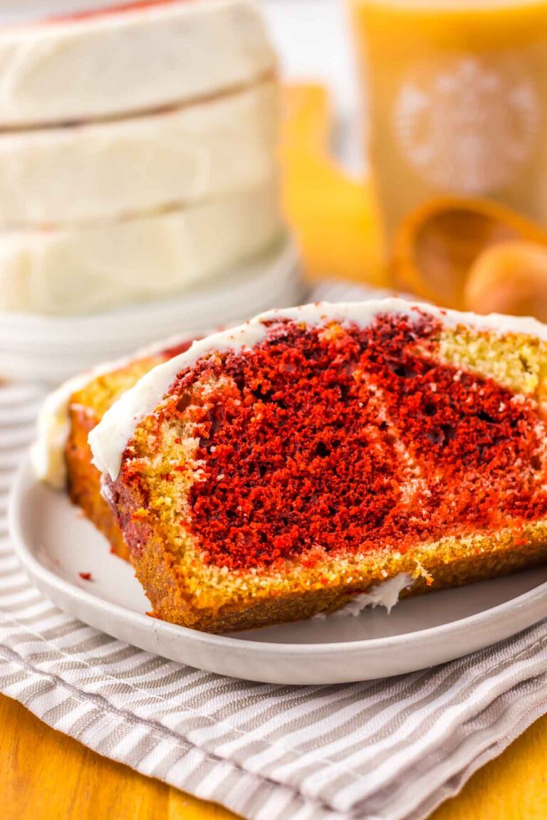 A slice of marbled red velvet and vanilla cake with cream cheese frosting rests on a plate atop a striped cloth. It's a deliciously tempting copycat Starbucks Red Velvet Loaf that promises to satisfy your sweet cravings.