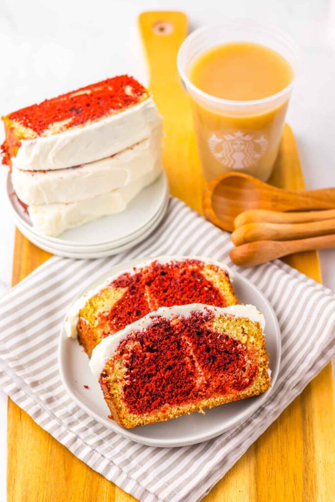 A copycat Starbucks Red Velvet Loaf and two slices of red velvet cake with cream cheese frosting sit invitingly on a plate beside a steaming cup of coffee, all elegantly arranged on a striped napkin with wooden spoons and a rustic wooden board.