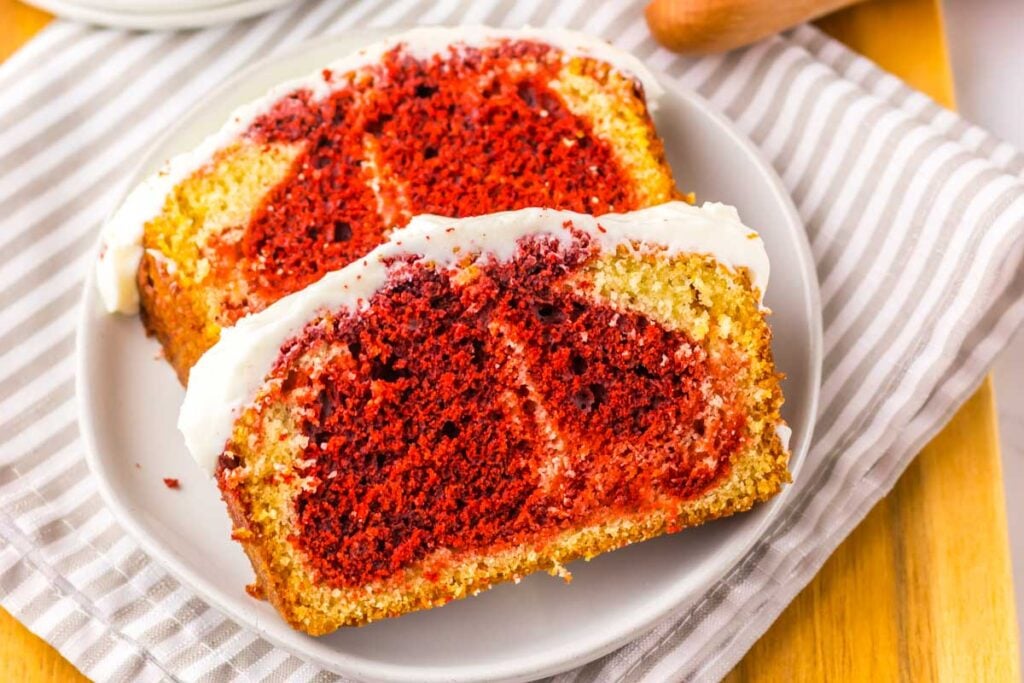 Two slices of Copycat Starbucks Red Velvet Loaf with red and yellow swirls, topped with white icing, are served on a white plate with a striped cloth underneath.