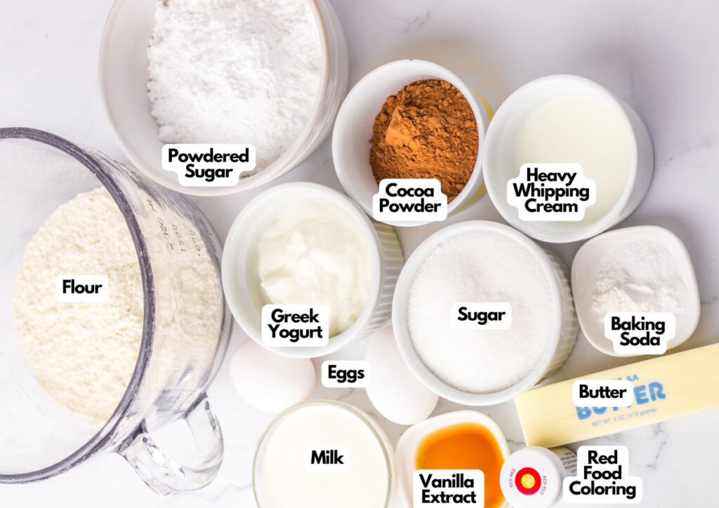 Various baking ingredients labeled in bowls and cups on a white surface, perfect for making a Copycat Starbucks Red Velvet Loaf—includes flour, cocoa powder, heavy whipping cream, Greek yogurt, eggs, butter, milk, vanilla extract, and red food coloring.