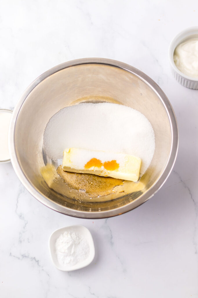 A mixing bowl brimming with butter, sugar, vanilla, and the familiar ingredients for a Copycat Starbucks Red Velvet Loaf stands ready for baking. Nearby, small bowls hold sour cream and baking powder to perfect this delectable treat.