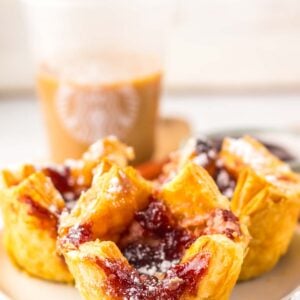 A close-up of copycat Starbucks Sugar Plum Danishes filled with jam and dusted with powdered sugar on a plate, paired perfectly with an iced coffee in the background.