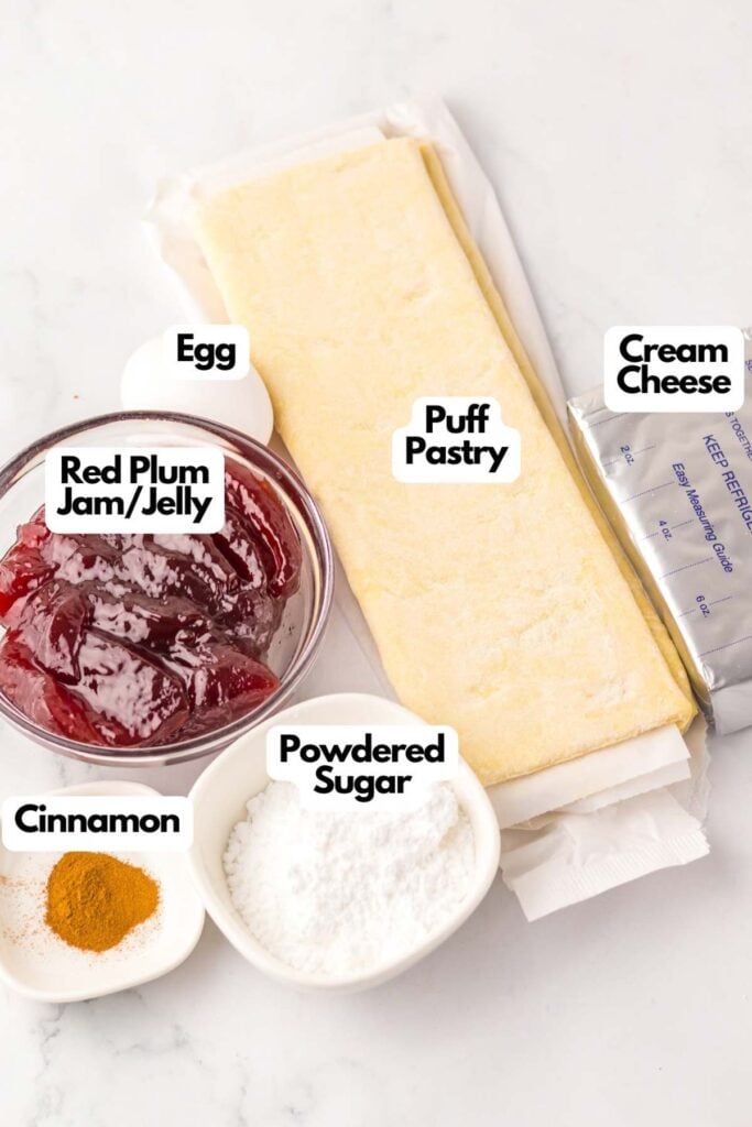 Ingredients for a copycat Starbucks Sugar Plum Danish recipe: puff pastry, cream cheese, red plum jam/jelly, egg, cinnamon, and powdered sugar neatly arranged on a surface.