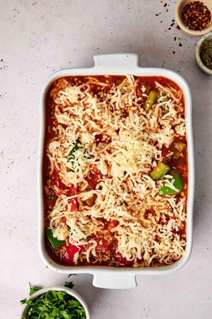 A stuffed pepper casserole dish filled with a layered mixture of cheese, tomato sauce, and vegetables, all topped with melted cheese. Various herbs and spices in small bowls sit beside the dish, enhancing its rich flavor profile.