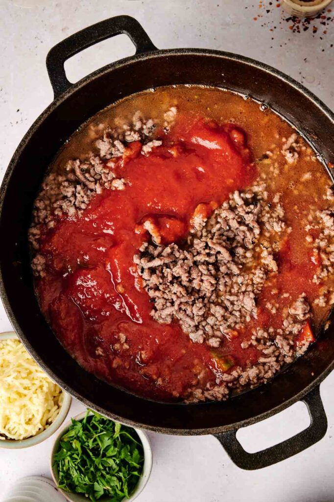 Ground beef and tomato sauce simmer in a black pot, surrounded by bowls of chopped herbs and grated cheese, setting the stage for a flavorful stuffed pepper casserole.
