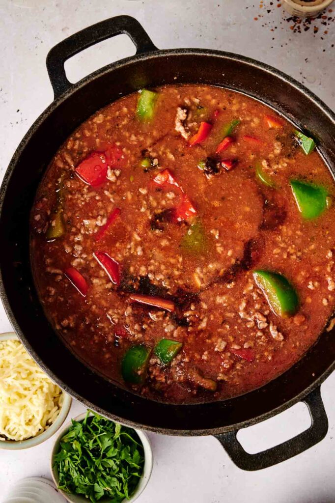 A pot of chili with ground meat and the flavors of a stuffed pepper casserole, featuring green and red bell peppers. Accompanied by small bowls of grated cheese and chopped herbs.