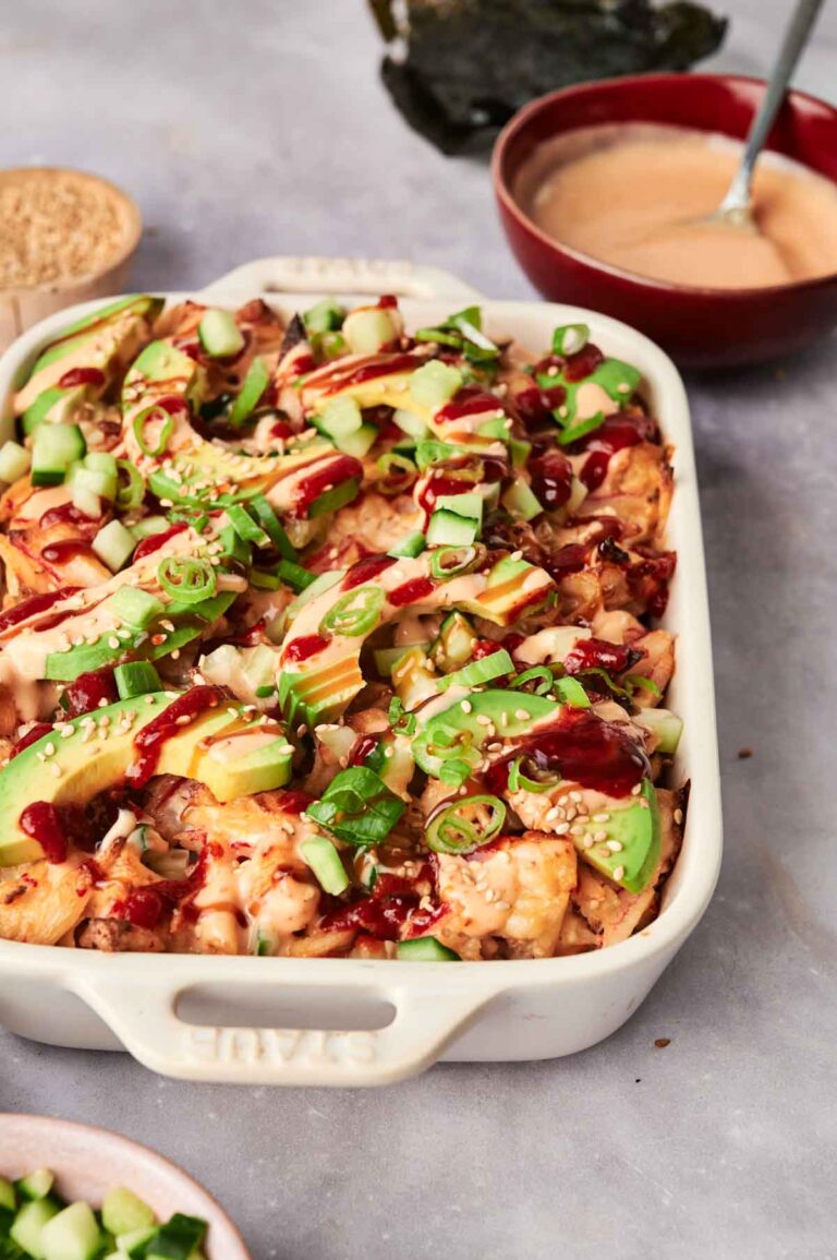 A casserole dish brimming with sushi bake is topped with avocado slices, green onions, and sesame seeds, all drizzled with sauce. In the background sits a bowl of dipping sauce, ready to accentuate the flavors.