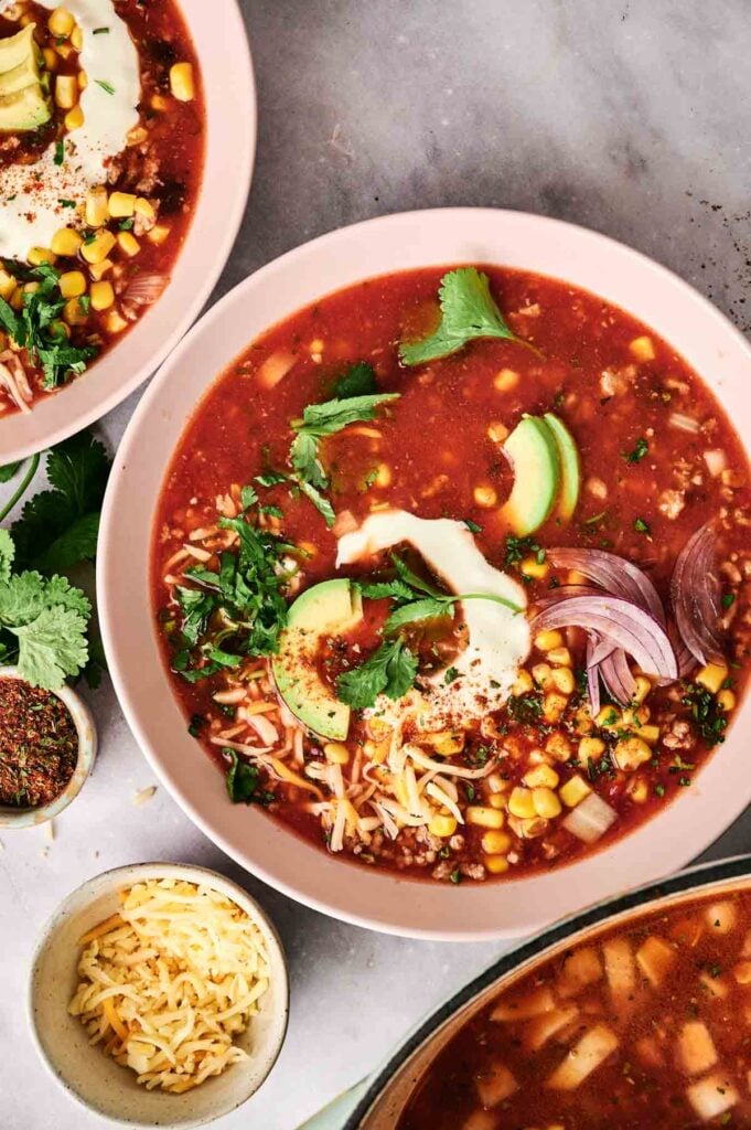 A bowl of taco soup garnished with avocado slices, shredded cheese, cilantro, corn, and red onion rests on a marble surface.