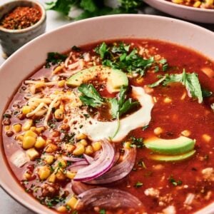 A bowl of taco soup rich with tomato flavor, topped with avocado slices, onion, corn, and herbs, finished with a dollop of cream. Another bowl and a dish of spices peek from the background.