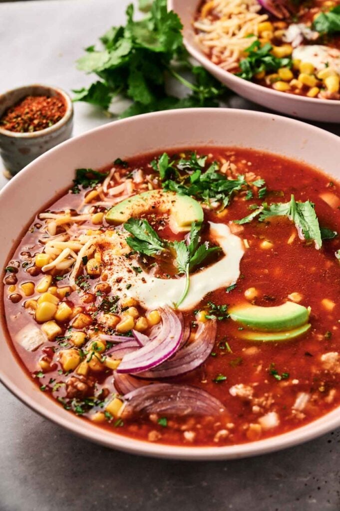 A bowl of taco soup rich with tomato flavor, topped with avocado slices, onion, corn, and herbs, finished with a dollop of cream. Another bowl and a dish of spices peek from the background.