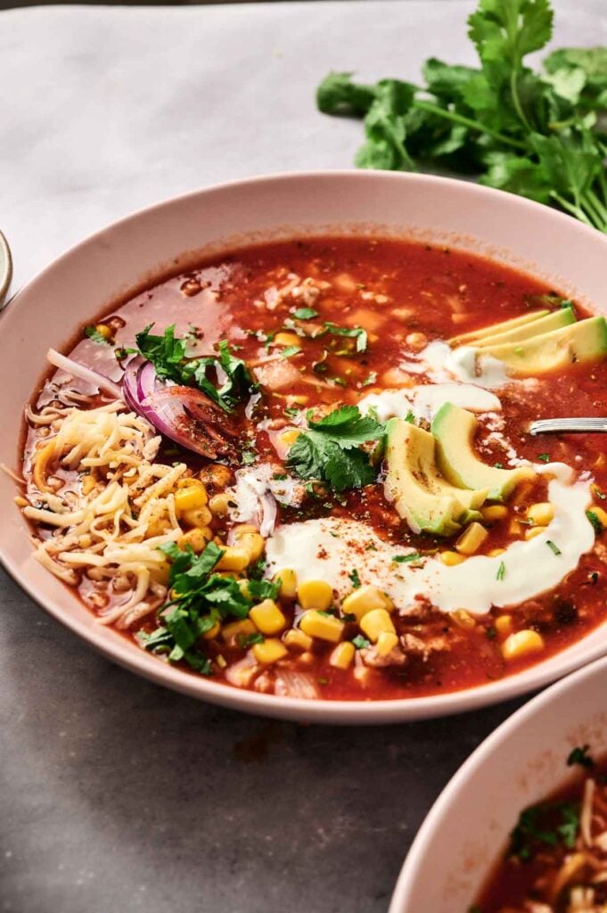 A bowl of taco soup brimming with a rich tomato base, adorned with avocado slices, corn, cheese, red onion, sour cream, and cilantro. A fresh sprig of cilantro rests beside the bowl.
