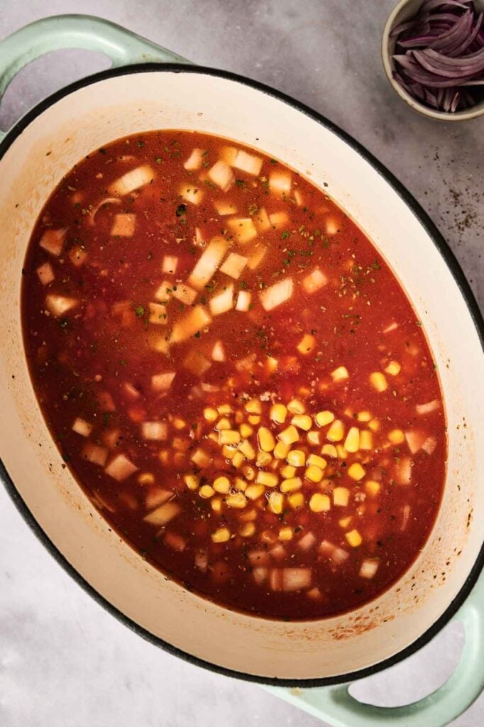 A pot of hearty taco soup with diced onions, corn, and herbs in a flavorful red broth. Bowl of sliced red onions in the background.