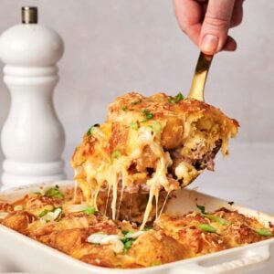 A hand lifts a cheesy tater tot breakfast casserole serving from a baking dish. A white pepper grinder is in the background.