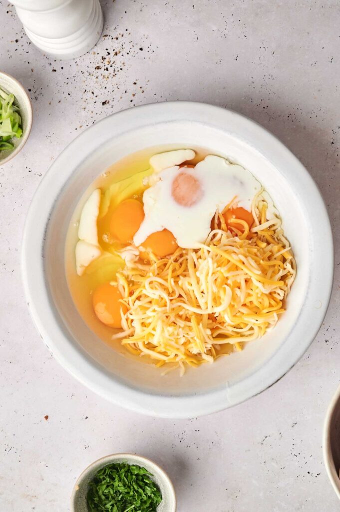 A white bowl containing shredded cheese, four cracked eggs, and cream sits on a light speckled surface, ready to be transformed into a tater tot breakfast casserole. It's surrounded by small bowls of green herbs and other ingredients.