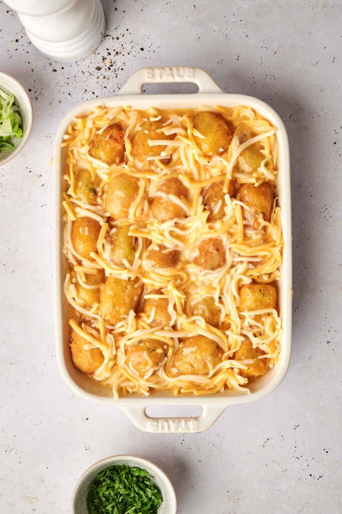 A tantalizing tater tot breakfast casserole topped with shredded cheese sits in a rectangular dish on a light-colored surface. Nearby, bowls of chopped green onions and parsley offer the perfect garnish to this morning delight.