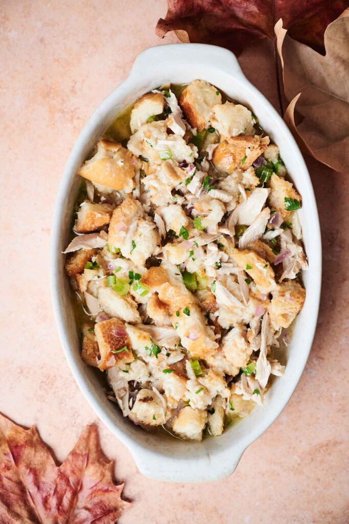 A baking dish filled with traditional stuffing, featuring bread cubes, turkey pieces, and garnished with herbs. Leaves are scattered around the dish on a peach-colored surface.