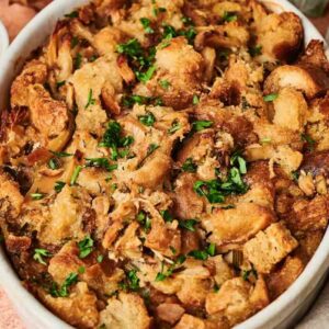 Oval dish of golden-brown stuffing garnished with chopped herbs. Bowl of herbs and autumn leaves nearby on a light surface.