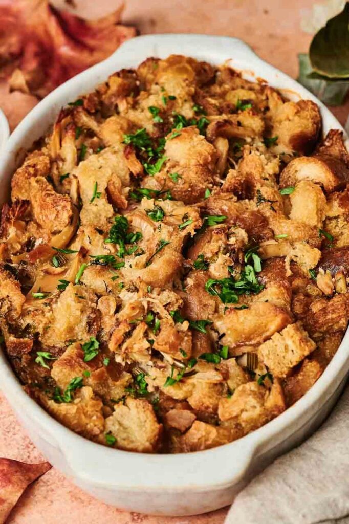 Oval dish of golden-brown stuffing garnished with chopped herbs. Bowl of herbs and autumn leaves nearby on a light surface.