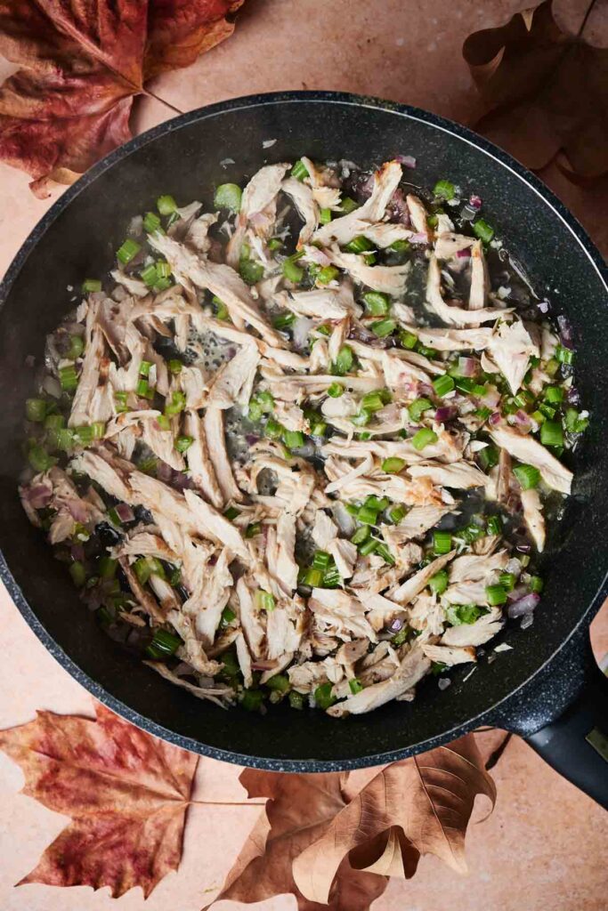 Cooked shredded chicken with diced green peppers and onions in a black frying pan, surrounded by autumn leaves on a countertop.