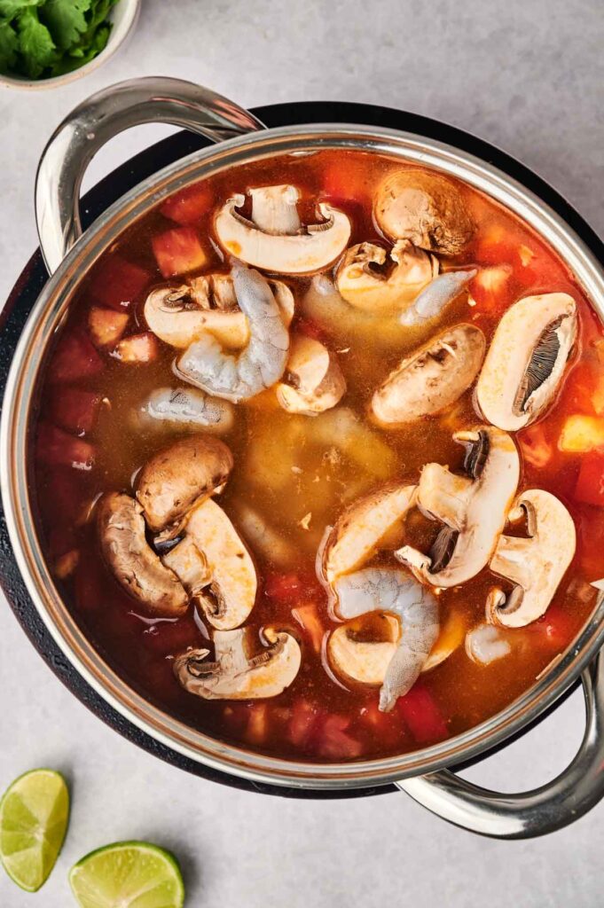 A pot of tom yum soup with sliced mushrooms and shrimp in a rich red broth. A lime wedge and fresh cilantro accompany this vibrant dish.