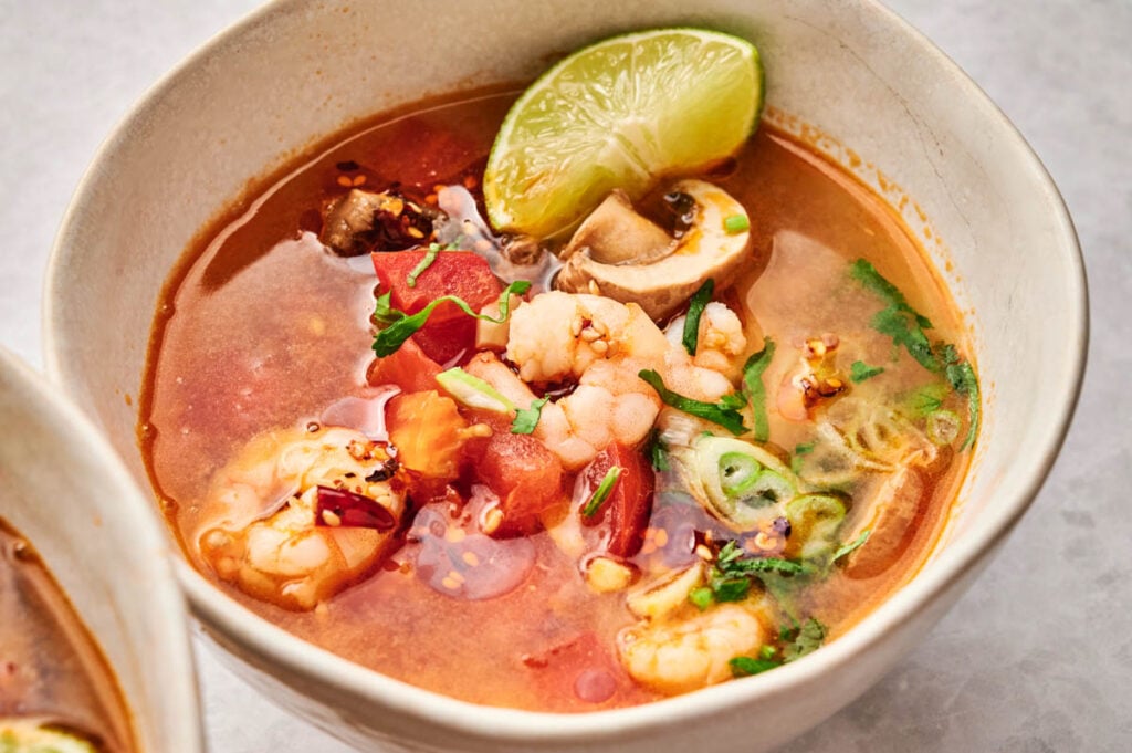 A bowl of tom yum soup with shrimp, mushrooms, tomato, herbs, red pepper flakes, and a lime wedge on the rim.