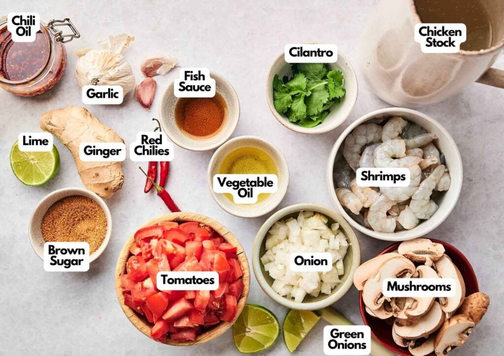 Top-down view of labeled ingredients for tom yum soup, featuring shrimp, tomatoes, onion, mushrooms, green onions, cilantro, fish sauce, chili oil, garlic, ginger, lime, brown sugar, chiles, and chicken stock.
