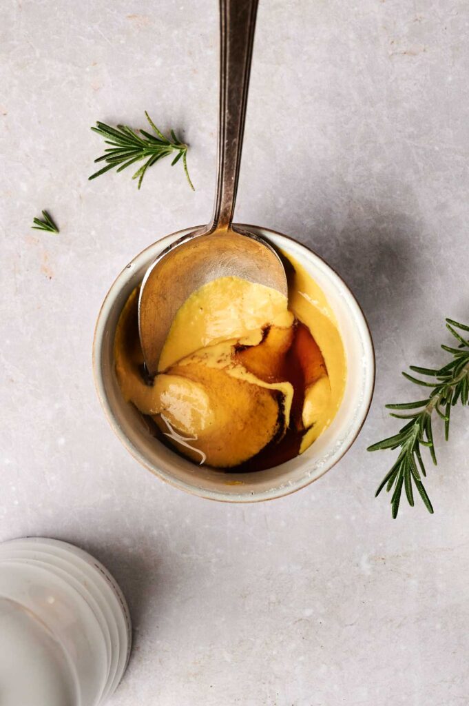 A spoon rests in a small bowl containing mustard and a dark liquid. Fresh rosemary sprigs are nearby on a light textured surface.
