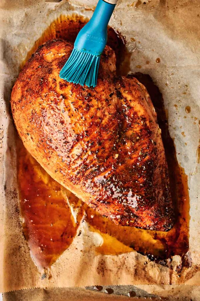 A roasted turkey breast on parchment paper is being brushed with a blue basting brush.