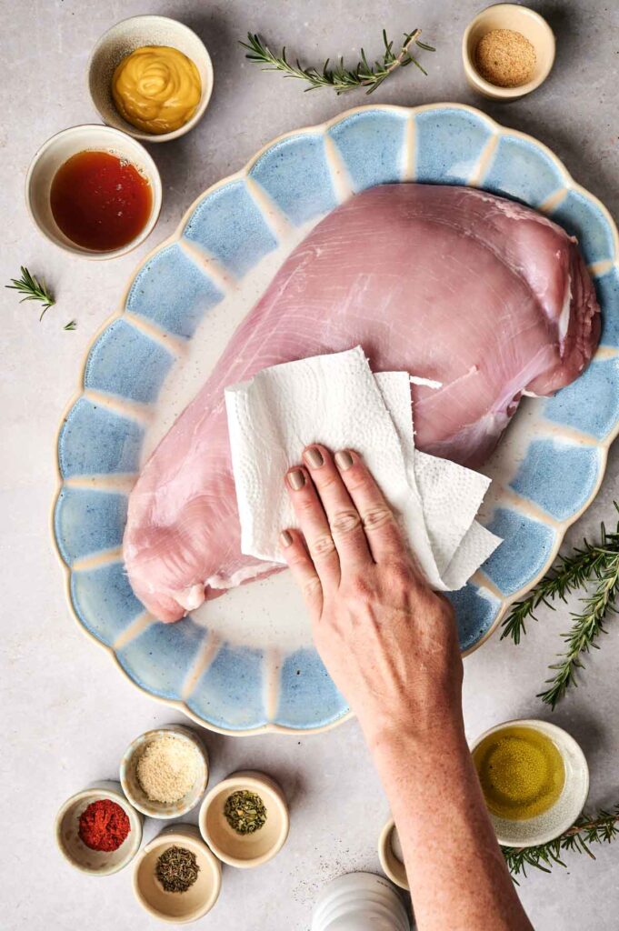 Raw turkey breast is being patted dry with a paper towel on a blue plate, surrounded by small bowls of various spices and ingredients.