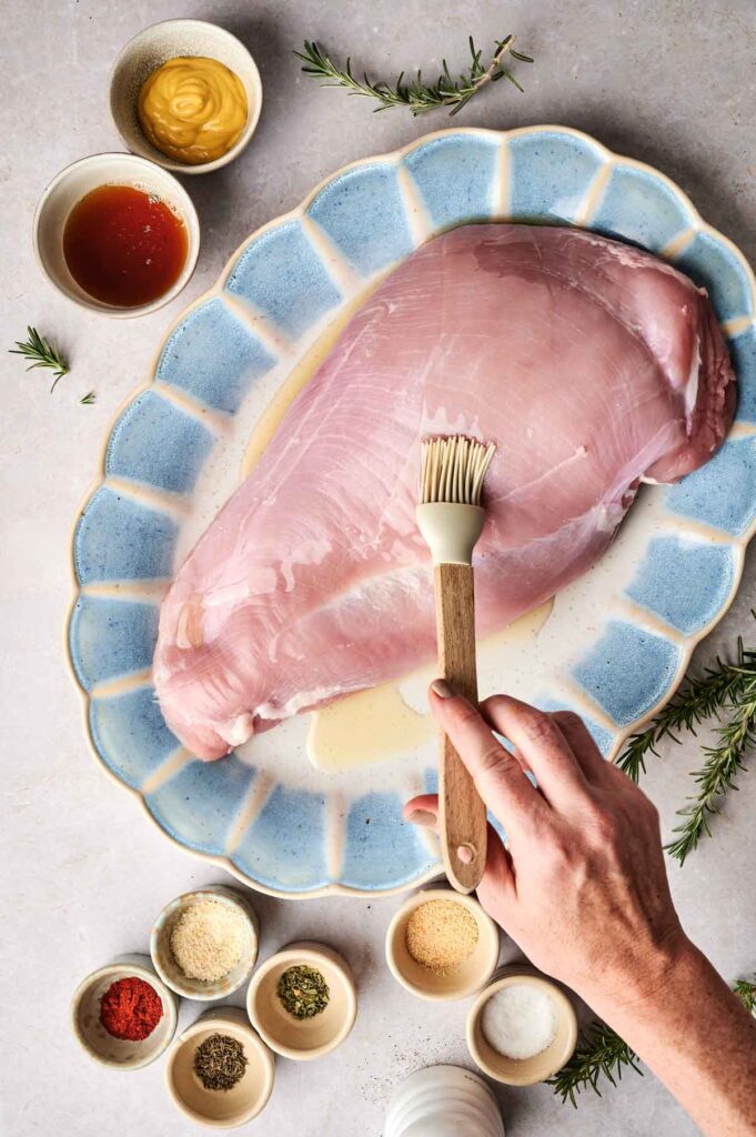 A hand uses a brush to apply marinade on a raw turkey breast. The turkey is placed on a blue platter, surrounded by small bowls of spices and herbs.
